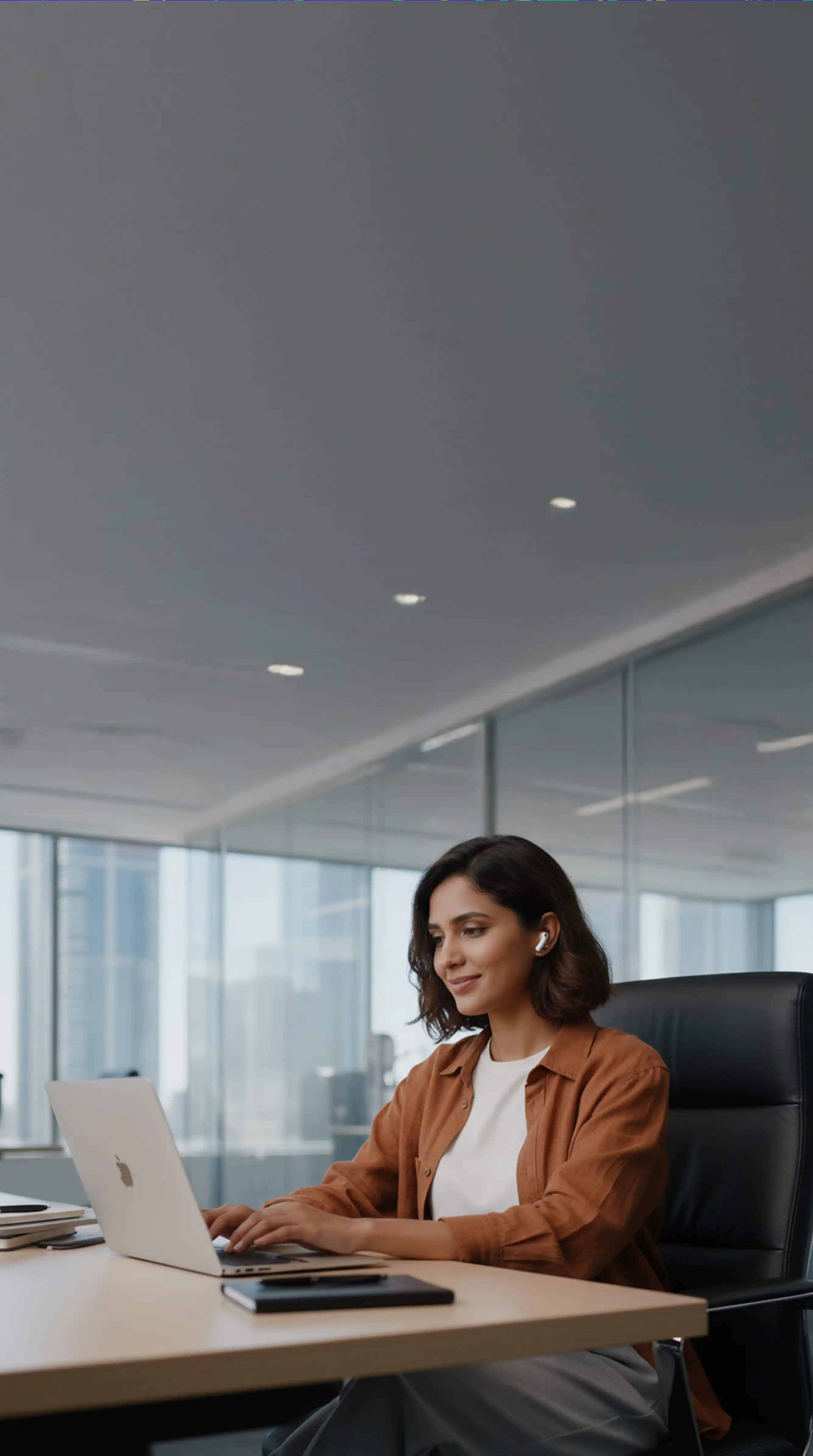 A woman is sitting her desk in a large office working on her laptop and wearing bluetooth earphones.