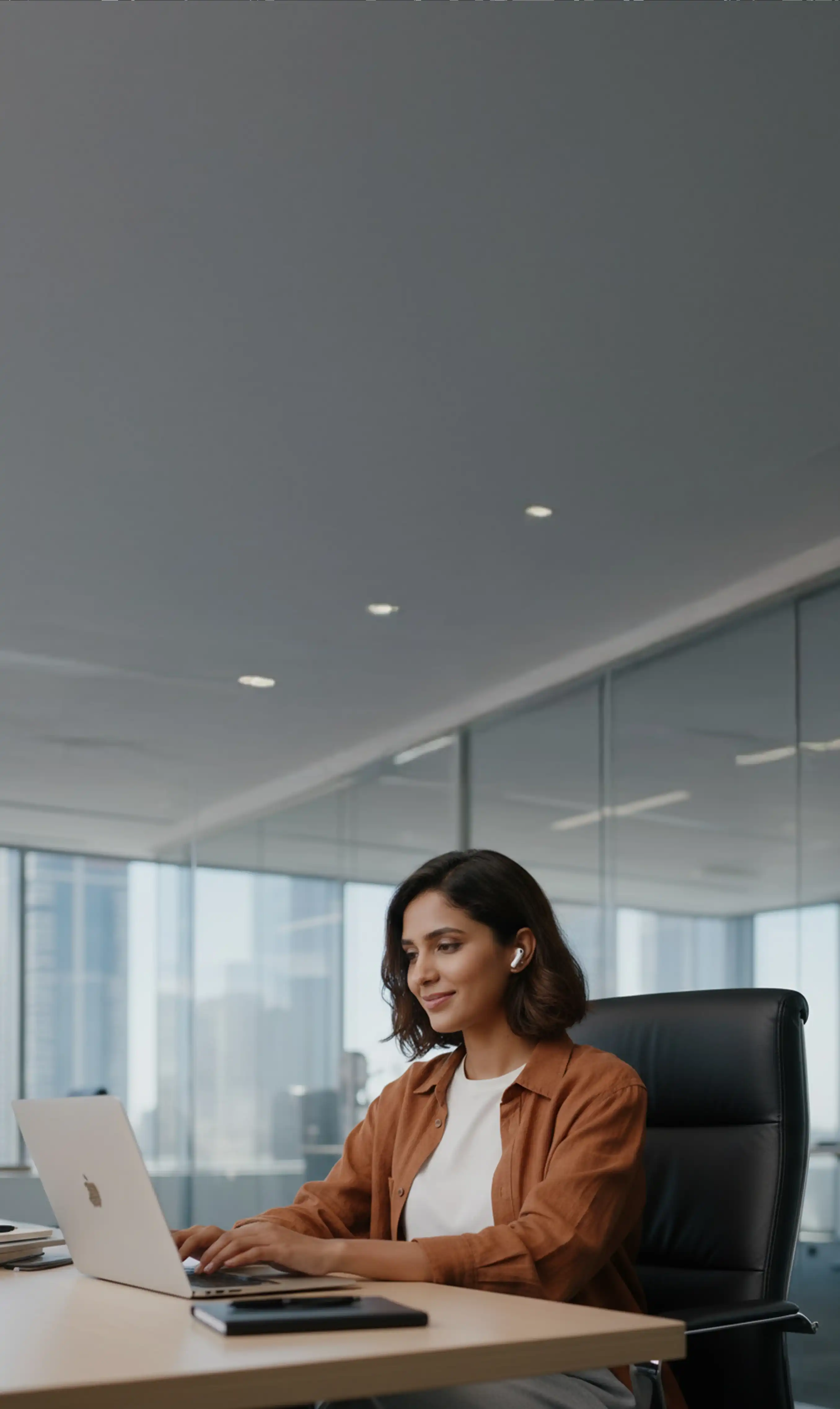 A woman is sitting her desk in a large office working on her laptop and wearing bluetooth earphones.