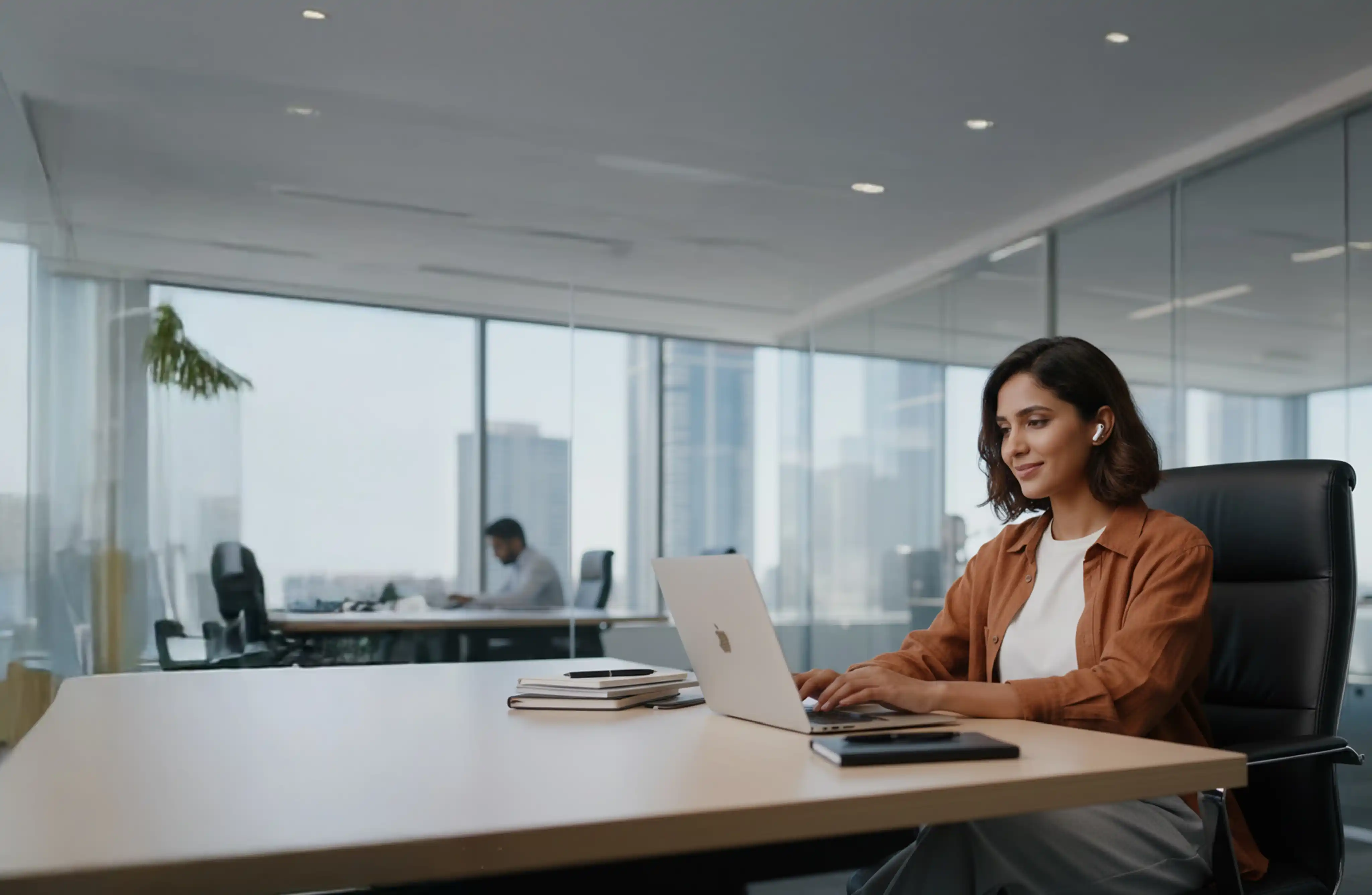 A woman is sitting her desk in a large office working on her laptop and wearing bluetooth earphones.