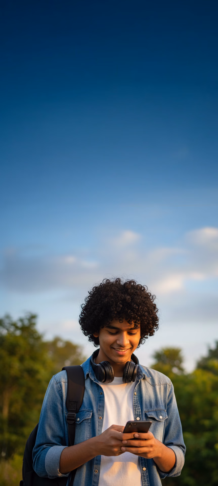 A teenage boy, standing in a park, smiling and looking in his phone.