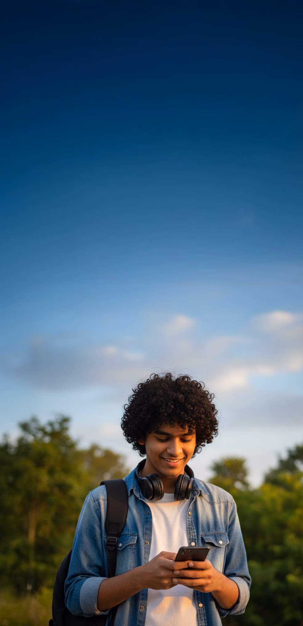 A teenage boy, standing in a park, smiling and looking in his phone.