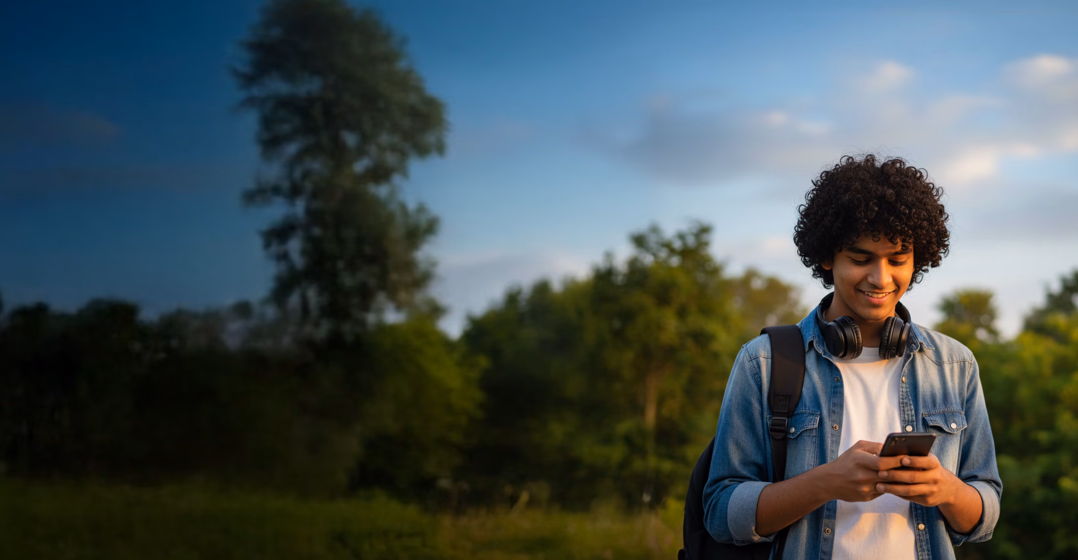 A teenage boy, standing in a park, smiling and looking in his phone.