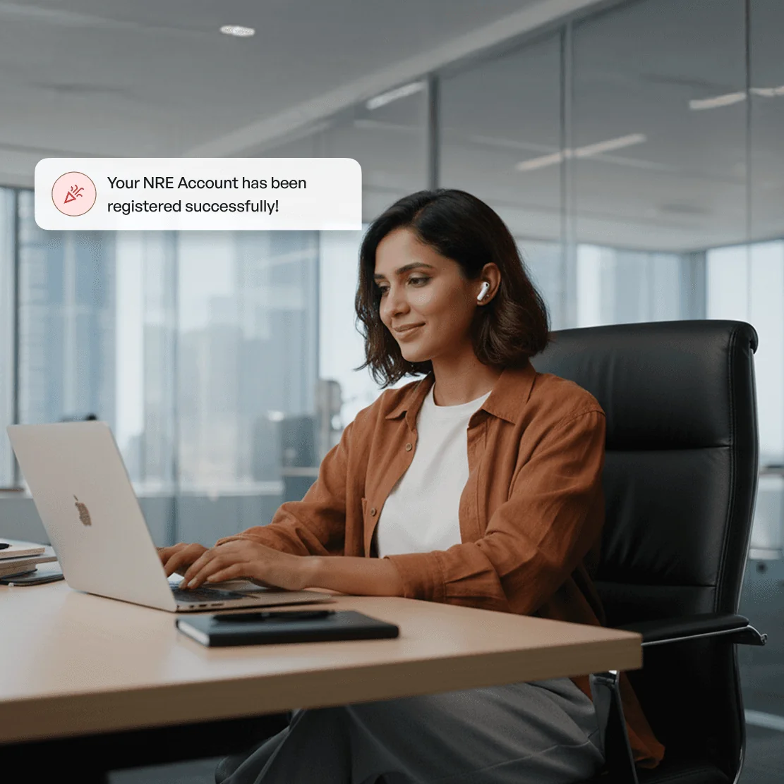 A woman is sitting her desk in a large office working on her laptop and wearing bluetooth earphones.